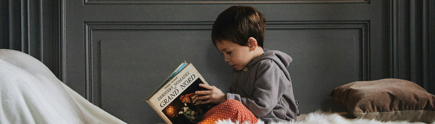 A boy in a grey hoodie sitting in bed and reading book.